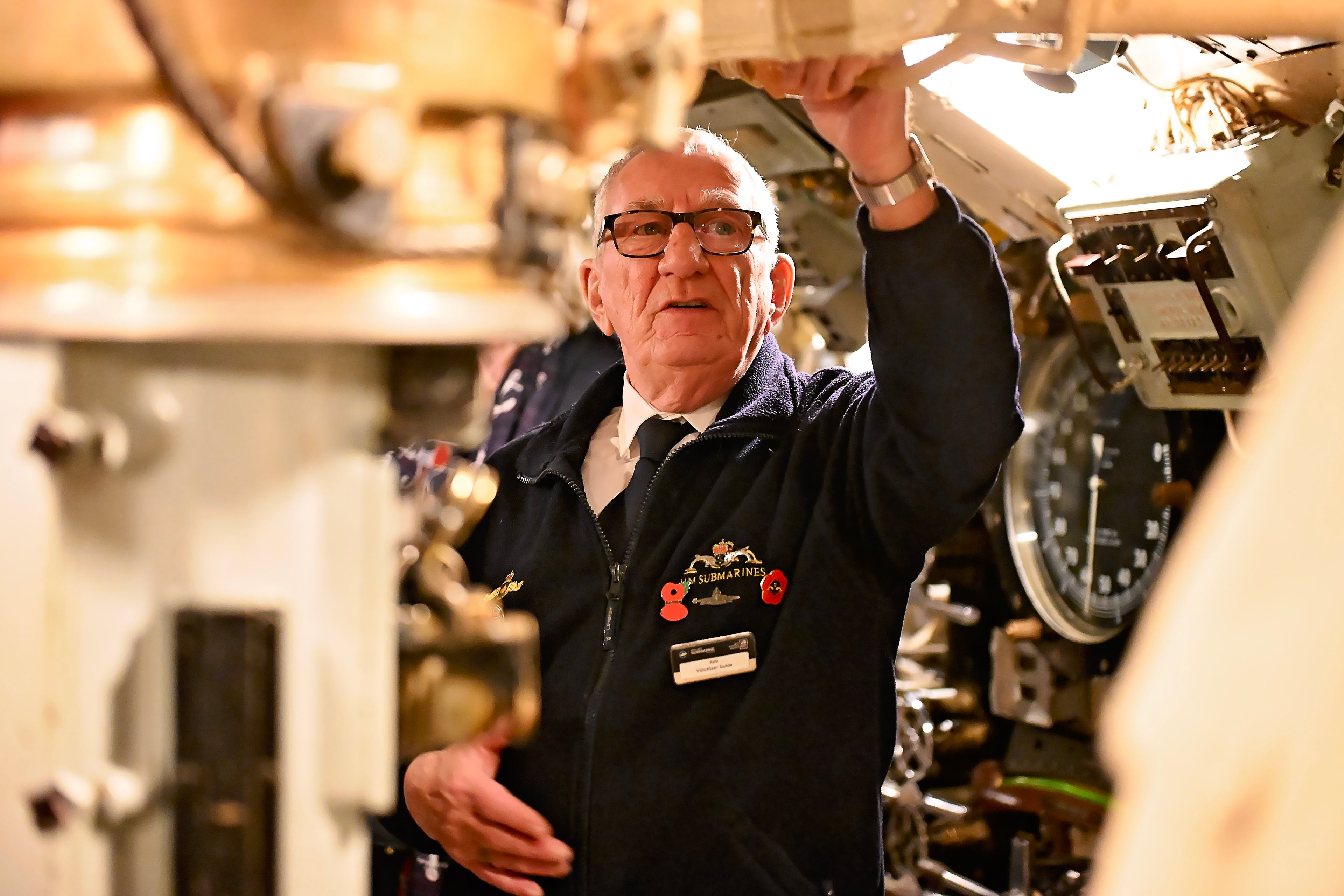 Man in a dark jacket with a name tag inside a submarine control room